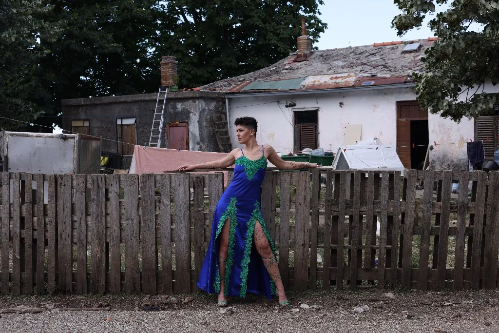Model in a cobalt gown with green lace against a rural fence and worn house