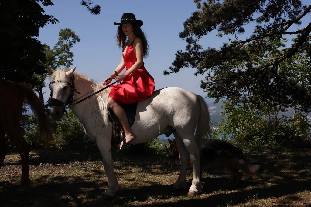Model in a red dress and dark hat on a white horse above the bay